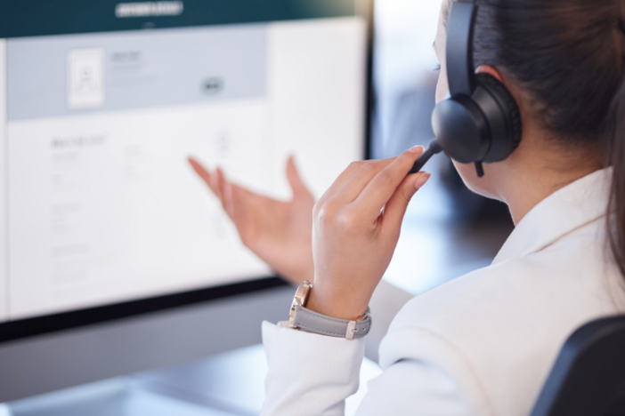 A woman wearing headphones speaks into a built-in microphone while looking intently at her computer screen.
