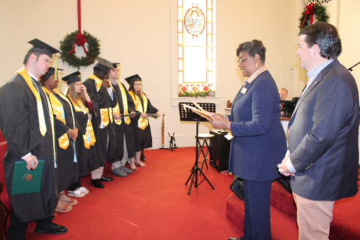 A group of graduates standing side by side standing in front of a woman reading from a document. 