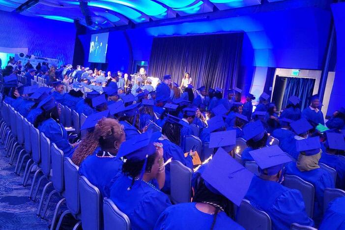 Graduates sitting down wearing blue graduation gown and cap watching the presenter speak. 