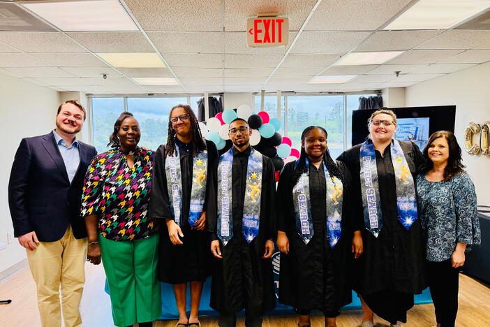 Seven people standing side by side and four of them are wearing graduation gown with a blue and black stole. 