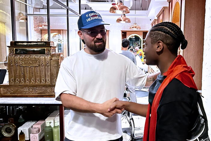 Two people shaking hands. One person wears a white t-shirt and a blue baseball cap. The other person wears a black t-shirt with an orange cloth draped over their shoulders and has braided hair.