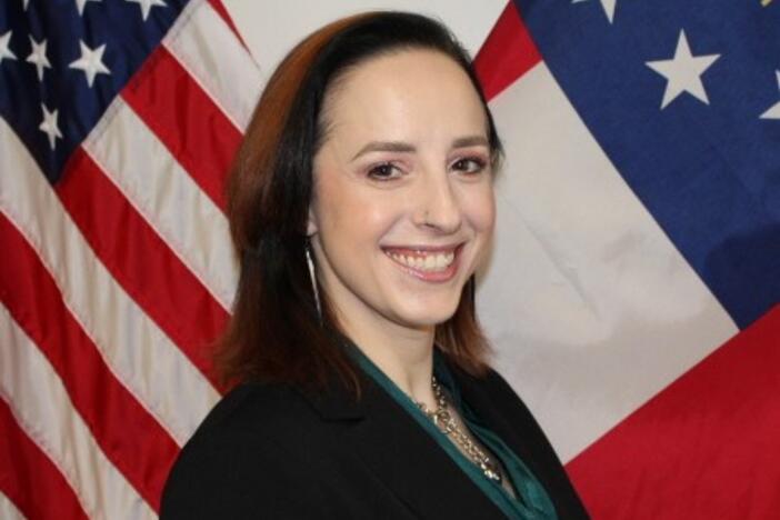 A woman in a black blazer and teal scarf smiles in front of the American flag and the Georgia state flag.