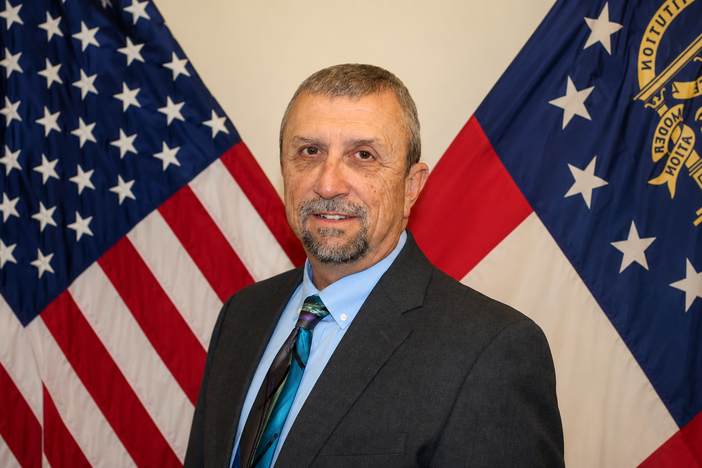 A man wearing a dark colored suit smiling behind the American and state flag. 