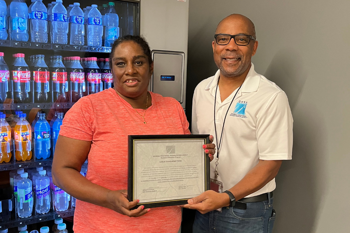 A person in an orange shirt and a person in a white polo shirt stand in front of a vending machine holding a framed certificate.