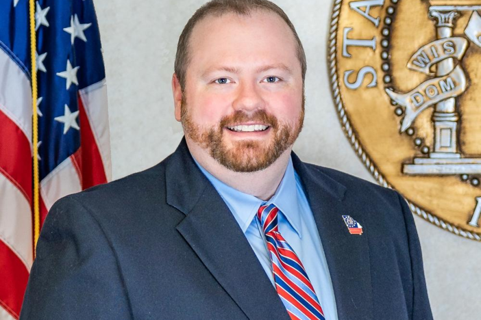 A man wearing a navy-blue suit standing behind an American flags. 