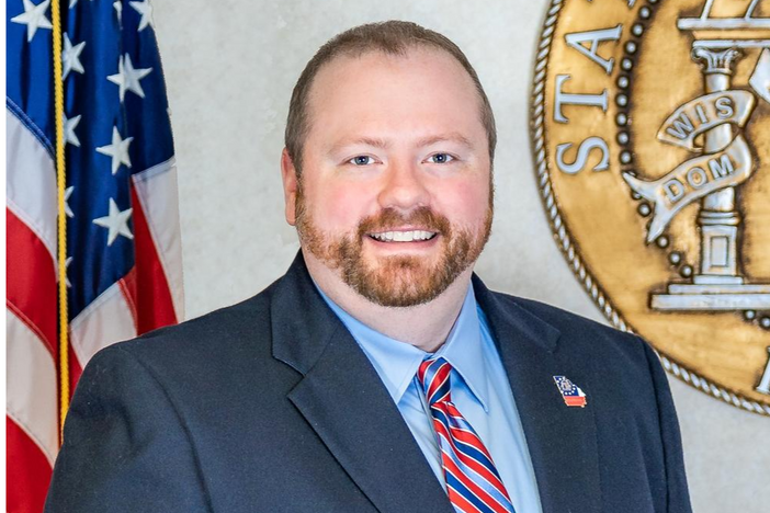 A man in a suit and tie smiles in front of an American flag and a state seal.