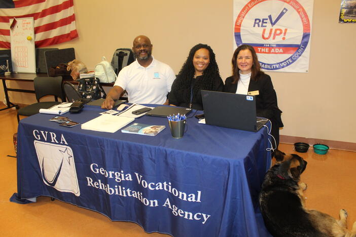 Three people seated behind a blue table at a community booth, with a laptop and brochures; the banner reads “Georgia Vocational Rehabilitation Agency,” and a dog sits on the floor in front.