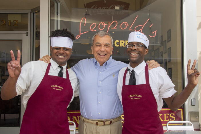 Two chefs in burgundy aprons and white headbands pose outside a restaurant entrance, smiling and making peace signs.